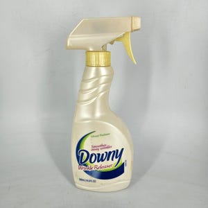 May include: A pearlescent white Downy Wrinkle Releaser spray bottle with a gold trigger and nozzle. The bottle has a curved design and features the Downy logo in blue and green, with the text "Wrinkle Releaser". The bottle contains 500ml (16.9 fl oz).