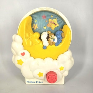 Puede incluir: Una luz de noche musical vintage de Fisher-Price con una luna creciente amarilla, nubes blancas y un fondo azul estrellado. Dos ositos de peluche están acurrucados en la luna. La luz de noche tiene una perilla roja y el logotipo de Fisher-Price.