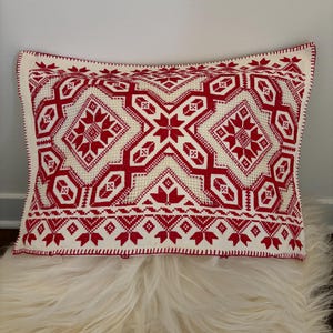 Könnte beinhalten: Rechteckiges Zierkissen mit weißem Hintergrund und rotem geometrischen Muster. Das Muster zeigt sternförmige und florale Motive. Das Kissen hat einen rot genähten Rand und liegt auf einer flauschigen, weißen Oberfläche.