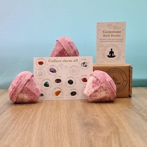 Könnte beinhalten: Drei rosafarbene Badebomben in Form von Edelsteinen, mit einer Karte, die eine Vielzahl von Kristallen zeigt und den Text "Collect them all". Die Badebomben befinden sich auf einer Holzoberfläche. Der Text "Gemstone Bath Bombs" befindet sich auf einer weißen Karte mit einer kleinen Illustration einer meditierenden Person.