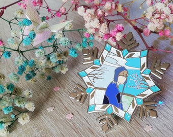 May include: A metal enamel pin shaped like a snowflake with a central illustration of a person wearing a blue hooded jacket. The pin features light blue and white accents, metallic silver outlines, and a winter-themed landscape background.