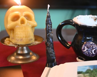 May include: A lit yellow skull-shaped candle sits on a gold stand, next to a black candle with a white tip, and a black ceramic mug with a white and purple moon and star design.