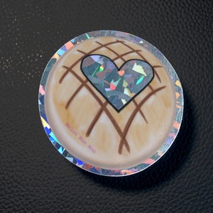 May include: A round sticker with a holographic finish. The sticker features a brown and white illustration of a pastry with a heart-shaped design. The text "Melon Bun Mom" is printed on the sticker.