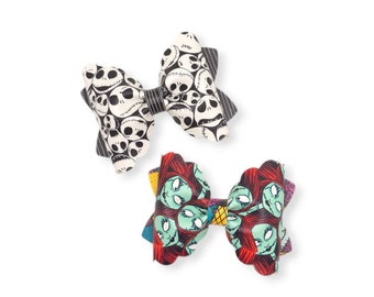 May include: Two hair bows with scalloped edges. The top bow features a black and white pattern of skull faces. The bottom bow displays a colorful repeating pattern of green-skinned characters with red hair, accented with yellow and purple details.