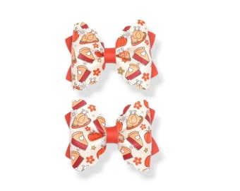 May include: Two hair bows with a Thanksgiving theme. The bows have a cream-colored fabric with a pattern of turkey, pumpkin, and pie slices. The center and the bow's tails are a solid red color. The bows are on a white background.