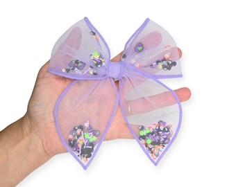 May include: A lavender tulle hair bow with a purple border. The bow is filled with colorful Halloween-themed sequins. The bow is held in a hand against a white background. The bow is a festive accessory.