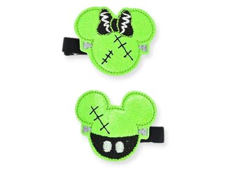 May include: Two green hair clips in the shape of a mouse head, with black stitched details and a black bow. The clips have a Frankenstein's monster theme, perfect for Halloween.