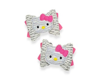 May include: Two hair bows featuring a Hello Kitty design. Each bow has a white and black striped pattern, a pink bow, and a silver Hello Kitty face with black eyes and a yellow nose. The bows are on a white background.