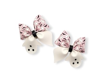 May include: Two Halloween-themed hair bows. Each bow features a pink fabric with white ghost and bat patterns, a white bow, and a ghost-shaped cutout with three black dots. The bows are connected by a black band.