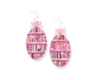 May include: Pink oval earrings with a bookshelf design featuring books and flowers. The earrings have a pink bow at the top.
