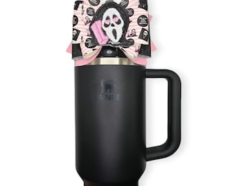 May include: A black Stanley tumbler with a black handle, topped with a decorative bow. The bow features a Scream mask design with pink accents and the text "Do you like scary movies?". The tumbler is designed to hold beverages.