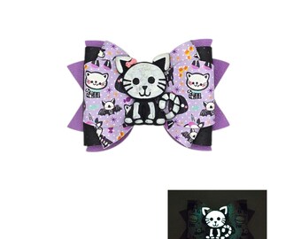 May include: A Halloween-themed hair bow featuring a black and white skeleton cat. The bow has a purple and black layered design with a repeating pattern of bats, cats, and unicorns. The bow glows in the dark.