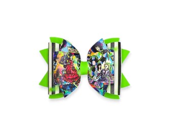 May include: A vibrant hair bow featuring a colorful graphic print with characters and the word "GIRLS" from the movie Beetlejuice. The bow has a neon green center and accents, with black and white striped edges.