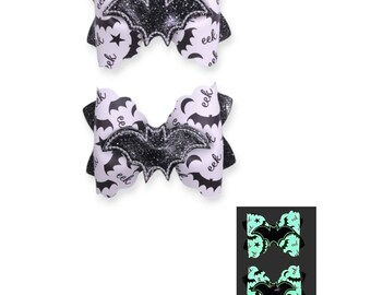 May include: Two hair bows featuring a white background with a black pattern of bats, stars, moons, and the text eek. Each bow is centered with a black glitter bat applique. A small inset shows the bows glowing in a light green color in the dark.