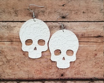 May include: A pair of white skull-shaped earrings featuring a delicate lace-like embossed texture. Each skull has two circular eye cutouts and a triangular nose opening. The earrings are attached to silver-toned metal fishhook ear wires.