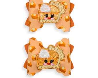 May include: Two orange hair bows with a pumpkin pie design. The bows have a repeating pattern of pumpkin pie slices and a pumpkin pie applique with a smiling face. The bows are a festive accessory.