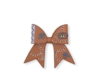 May include: A brown faux leather hair bow featuring embroidered white stitch patterns, a single green eye detail, and grey key motifs. The bow has a silver wavy edge on the left side and a central knot, with two pointed tails extending downwards.