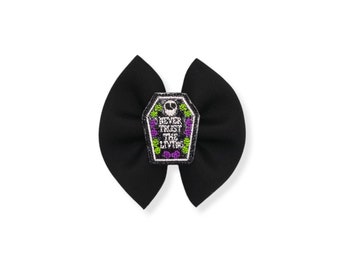 May include: A black fabric hair bow featuring a central embroidered patch shaped like a coffin. The patch displays a white skull icon above the text NEVER TRUST THE LIVING, surrounded by small purple and green floral accents on a black background.