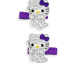 May include: Two Halloween-themed hair clips featuring a mummy design. Each clip has a white mummy character with a purple bow and is attached to a purple ribbon clip. The clips are designed for children.