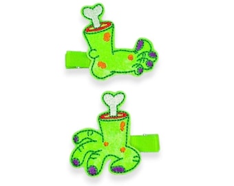 May include: Two green hair clips shaped like cartoon zombie feet. The feet are lime green with purple accents and orange spots. Each clip has a green ribbon base and a bone sticking out of the top of the foot.