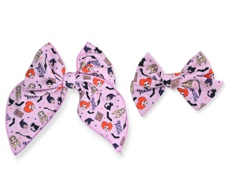 May include: Two light purple hair bows with a repeating pattern of cartoon witches, cats, and the words "Hocus Pocus". The bows are different sizes, with the larger one having long, flowing ends.