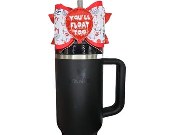 May include: A black Stanley tumbler with a handle, topped with a red and white bow. The bow features a repeating pattern of a clown and the text "YOU'LL FLOAT TOO". The tumbler has the Stanley logo.