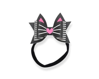 May include: A black cat-themed bow headband with a pink heart and ear accents. The bow features white stripes and is attached to a black elastic band. This accessory is perfect for adding a touch of whimsy to any outfit.