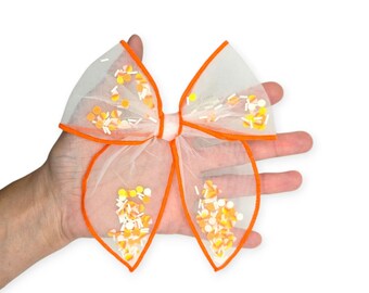 May include: A white tulle hair bow with orange trim and filled with candy corn-shaped confetti. The bow is held in a hand against a white background. The bow is a festive accessory.