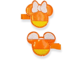 May include: Two orange and yellow hair clips in the shape of a mouse head with a white bow. The clips are designed to resemble candy corn. The clips are made of fabric and have an orange barrette clip.
