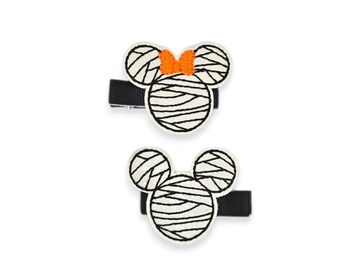 May include: Two hair clips featuring embroidered mouse-shaped icons wrapped in white bandages with black outlines. The top clip includes a small orange bow between the ears, while the bottom clip features the same mummy-style design without a bow.