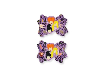 May include: Two hair bows featuring a purple and black leopard print pattern. Each bow is adorned with an embroidered patch depicting three figures with orange, blonde, and black hair. The text Hocus Pocus is visible on the fabric of the bows.