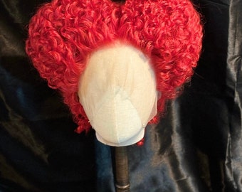 May include: A red curly wig styled in a heart shape. The wig is on a mannequin head and is displayed against a black background.