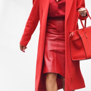 Op de afbeelding: Een monochroom rode outfit met een lange jas, een overhemd met knopen en een leren rok. Een bijpassende rode handtas wordt gedragen. De outfit wordt gecompleteerd met rode schoenen.