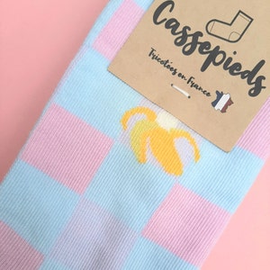 Può includere: Un paio di calzini con un motivo a quadri blu chiaro e rosa. Una banana gialla è stampata sui calzini. I calzini hanno un'etichetta che dice "Cassepieds" e "Tricotées en France" con una piccola bandiera francese.