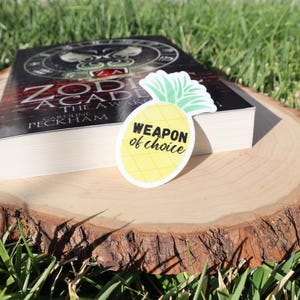 May include: A yellow pineapple sticker with the text "Weapon of choice" on a wooden surface with a book titled "Zodiac Academy: The Awakened" by Caroline Peckham.