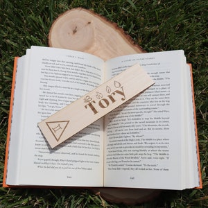 May include: A wooden bookmark with the name "Tory" engraved on it. The bookmark is decorated with a Gemini zodiac symbol and a small leaf design. The bookmark is resting on a book with the title "A Court of Silver Flames".