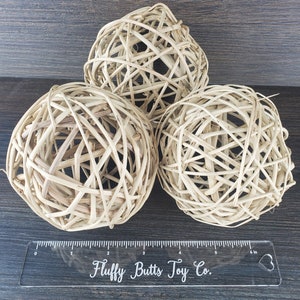 May include: Three natural woven wicker balls, approximately 6 inches in diameter, on a brown wooden surface.  The balls are made of natural woven straw and are a light beige color.  A ruler with the text "Fluffy Butts Toy Co." is in the foreground.