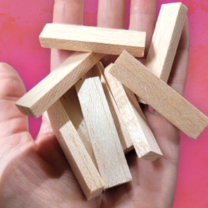 May include: A collection of small, rectangular wooden blocks, likely for crafts or building, are held in a hand. The blocks are a light, natural wood color and appear to be unfinished. The background is a solid pink.
