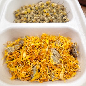 May include: A white container with two sections, each filled with dried flower petals. The top section contains small, light brown flower buds. The bottom section contains a large amount of yellow flower petals.