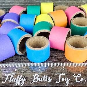 May include: A collection of colorful cardboard rings, each with a different color, including purple, blue, green, yellow, orange, and pink. The rings are stacked on top of each other and are arranged in a random order. The rings are all the same size and have a hole in the center. The rings are on a wooden surface with a ruler that says "Fluffy Butts Toy Co."
