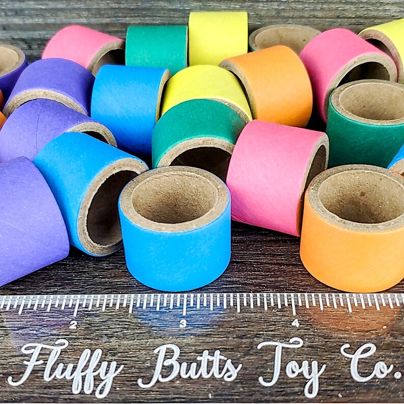 May include: A collection of colorful cardboard rings, each with a different color, including purple, blue, green, yellow, orange, and pink. The rings are stacked on top of each other and are arranged in a random order. The rings are all the same size and have a hole in the center. The rings are on a wooden surface with a ruler that says "Fluffy Butts Toy Co."