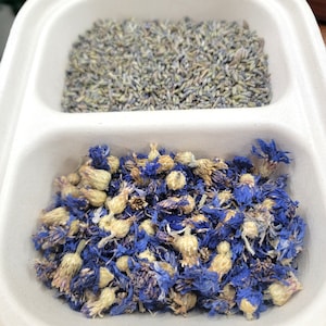 May include: A white container with two compartments. The top compartment contains dried lavender flowers. The bottom compartment contains dried blue cornflower petals.