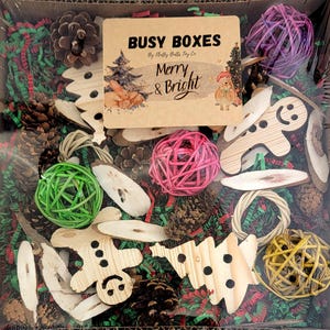 May include: A Christmas-themed busy box for pets, filled with natural and wooden toys. The box includes pine cones, wooden trees, a gingerbread man, and colorful woven balls. The box is decorated with red and green shredded paper and a sign that reads "Busy Boxes by Holly Bells Toy Co. Merry & Bright".