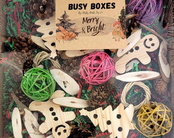 May include: A Christmas-themed busy box for pets, filled with natural and wooden toys. The box includes pine cones, wooden trees, a gingerbread man, and colorful woven balls. The box is decorated with red and green shredded paper and a sign that reads "Busy Boxes by Holly Bells Toy Co. Merry & Bright".