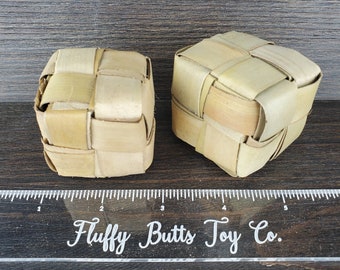 May include: Two woven, cube-shaped toys made from light brown, dried leaves. The toys are placed on a dark wood surface. A clear ruler with the text "Fluffy Butts Toy Co." is in the foreground.