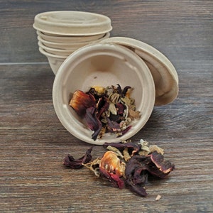 May include: A close-up of a brown paper bowl filled with dried hibiscus flowers, rose petals, and other herbs. The bowl is sitting on a wooden surface.