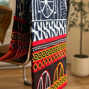 May include: A rectangular fabric textile featuring a dark blue and white geometric pattern at the top, followed by a horizontal red band with yellow and black decorative motifs, and a black section at the bottom with yellow arch-shaped designs.