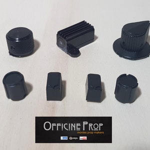 Peut inclure: Un ensemble de sept boutons en plastique noir, dont un bouton rond avec une surface texturée, un bouton rectangulaire et un bouton avec un sommet pointu. Les boutons sont disposés sur une surface brun clair.