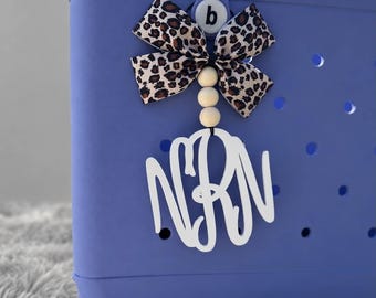 May include: A periwinkle blue perforated tote bag featuring a decorative charm attached to the handle. The charm consists of a leopard print fabric bow, three light-coloured wooden beads, and a white cutout monogram featuring the letters NRN.