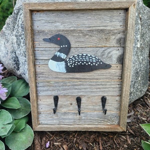 May include: A rustic wooden key holder with a painted loon design and three black metal hooks. The wood is weathered and has a gray finish.